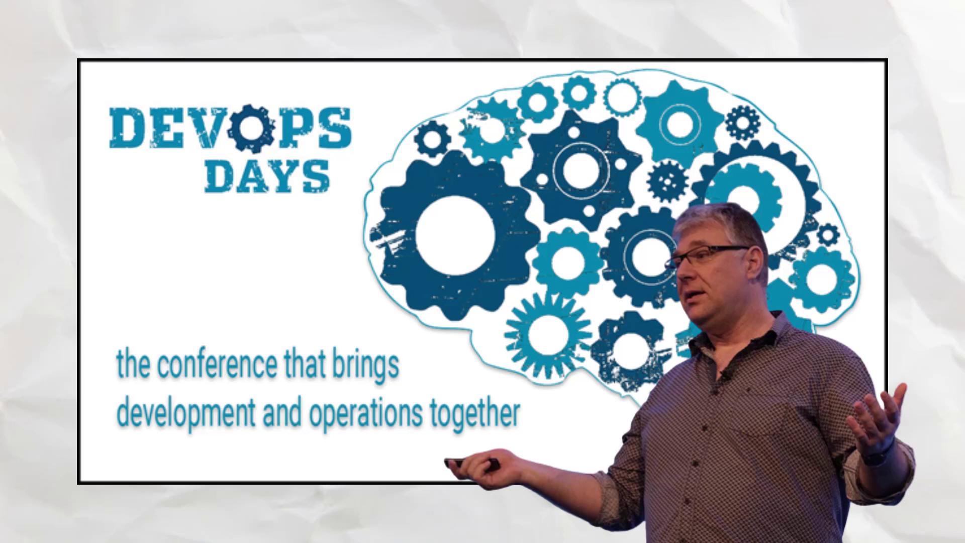 A speaker presents at a DevOps Days conference, featuring a brain-shaped gear design and the tagline "the conference that brings development and operations together."