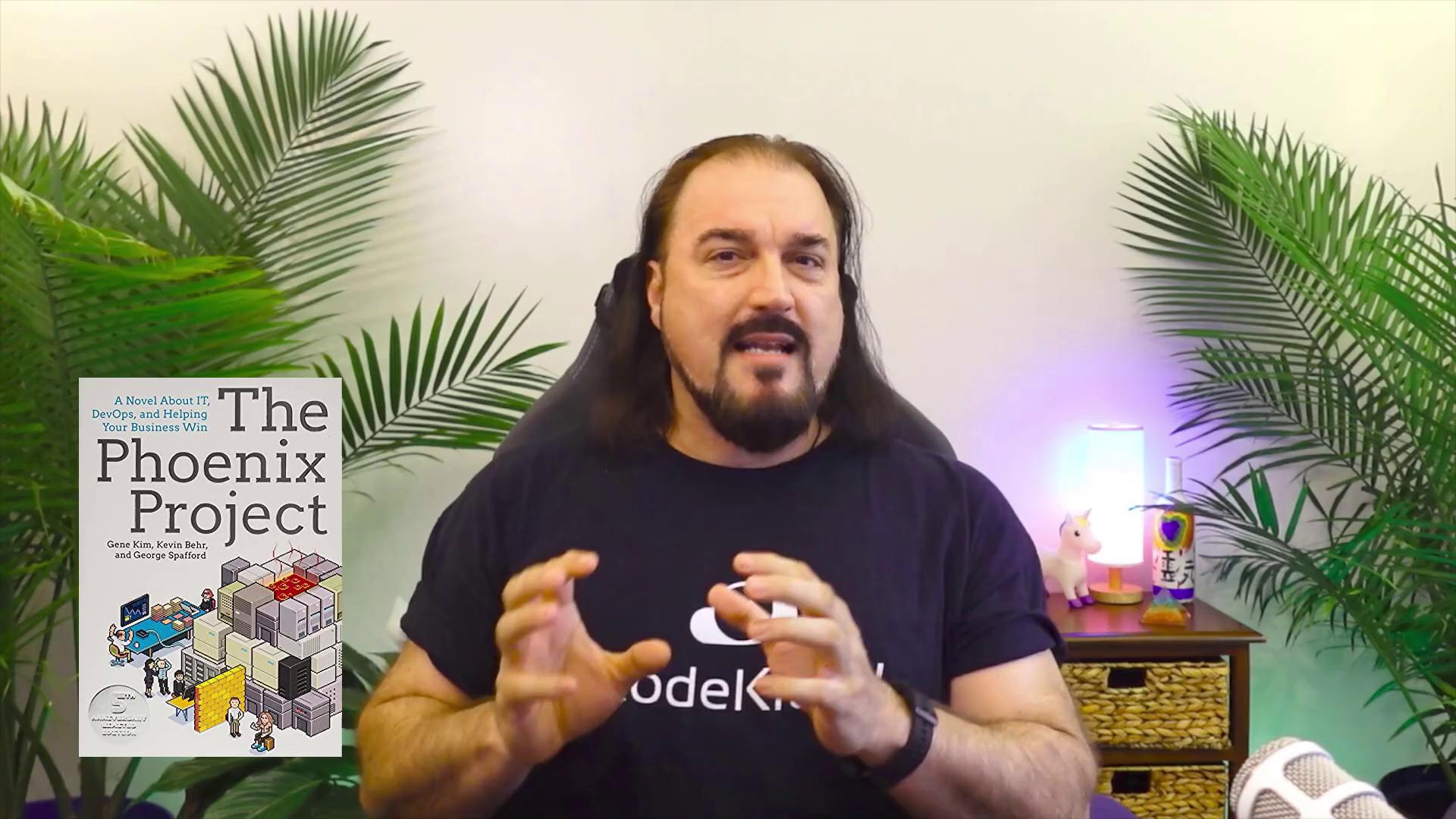 A person is speaking in front of a backdrop with plants, a book titled "The Phoenix Project," and a small lamp on a table.